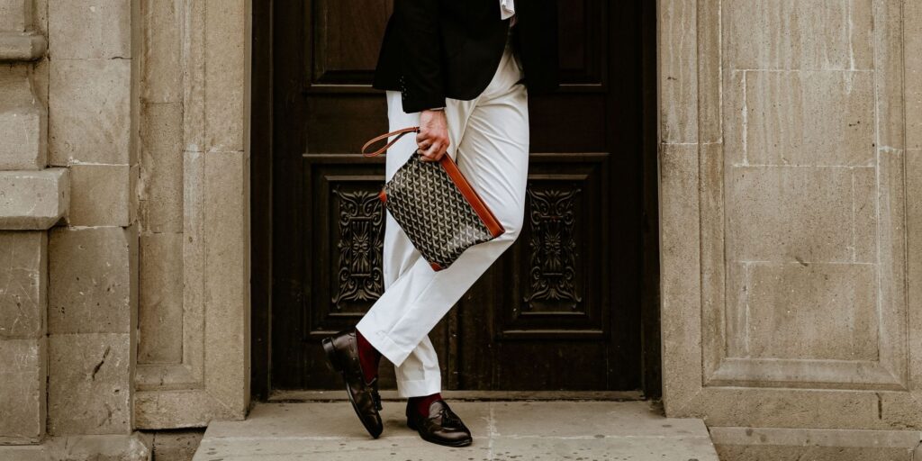 man holding a goyard bag. He is wearing white pants and leaning on a wall