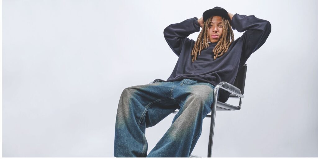 Man with dreadlocks leaning back in a chair. He is wearing jeans, a jumper and a hat from boohoo