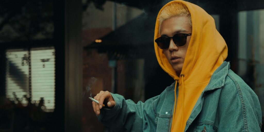 Asian man with bleached hair is smoking a cigarette and wearing a yellow jumper and green jacket