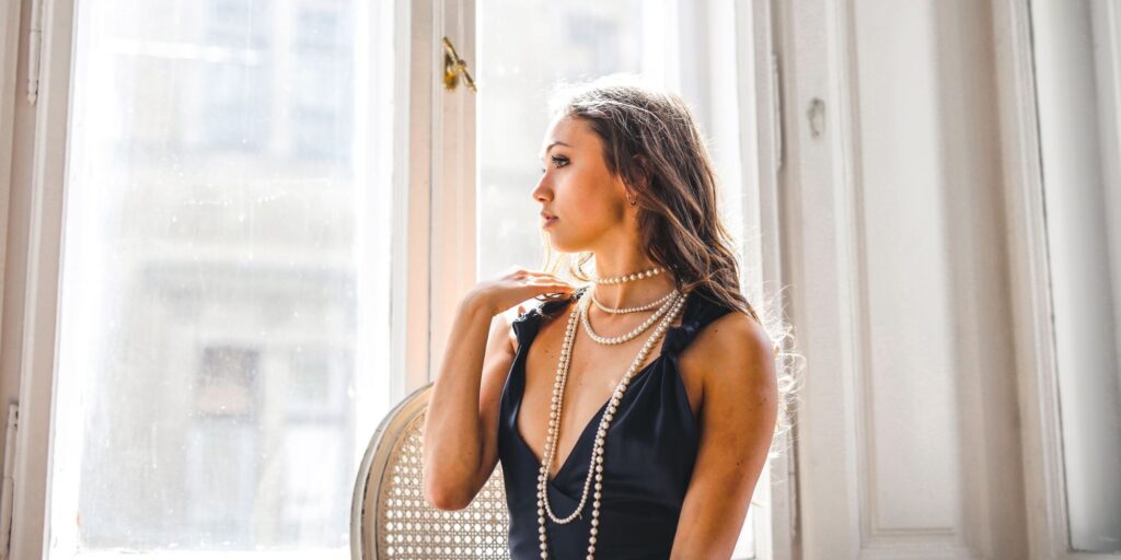 A woman looking out a window wearing a black dress and many pearl necklaces