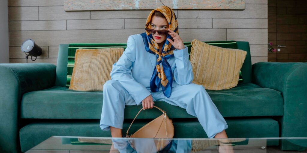 A woman sat on a green couch. She is wearing a blue suit set with a headscarf and sunglasses. She's very retro