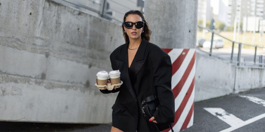 Woman wearing an oversized black blazer is walking in the city holding 2 coffees