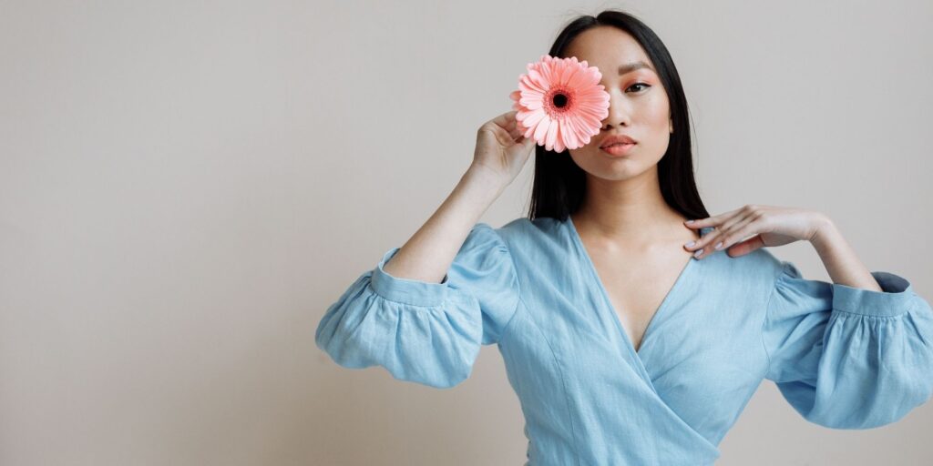 Beautiful Asian woman in a light blue dress holding a pink flower over one eye